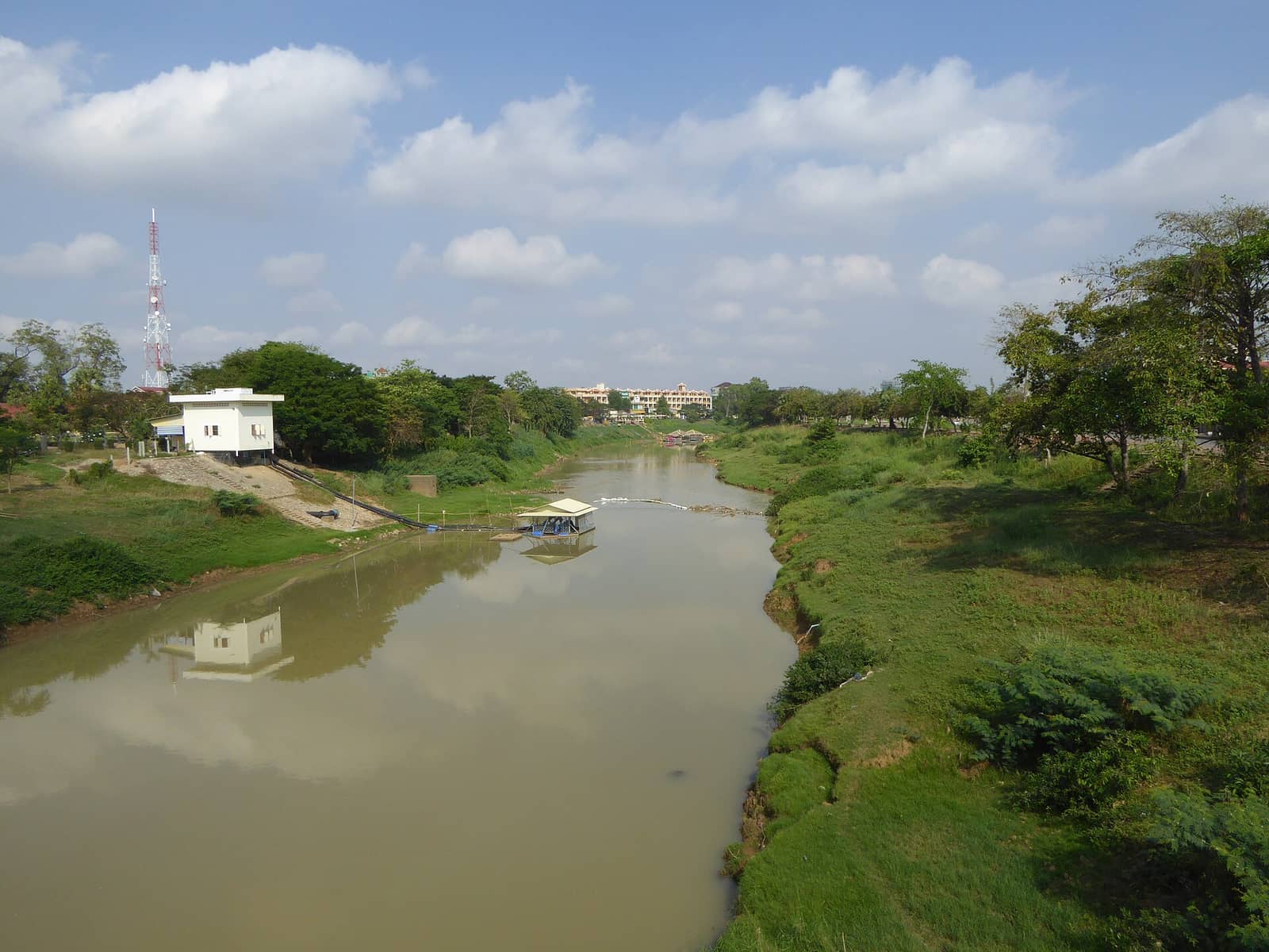 The Sangker River running through Battambang between grassy banks, with the town’s low buildings along the far side under a blue sky
