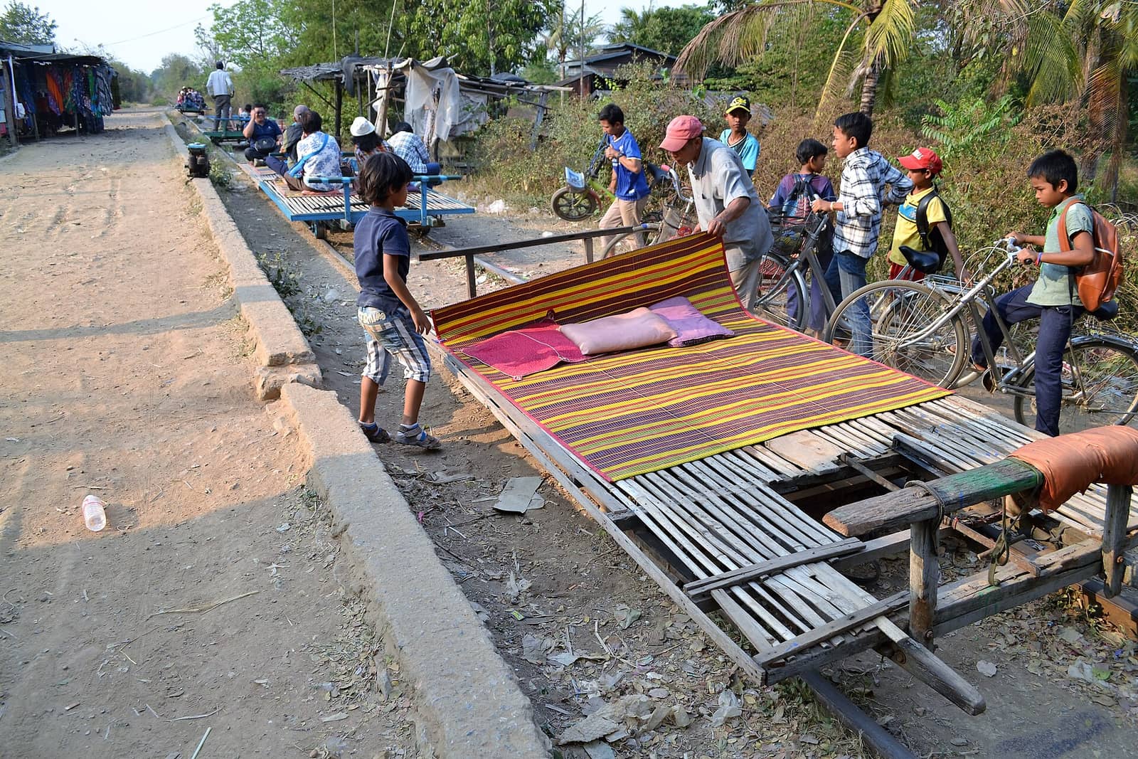 A bamboo norry — a slatted platform on rail wheels, spread with a striped mat and cushions — being lifted onto the track outside Battambang