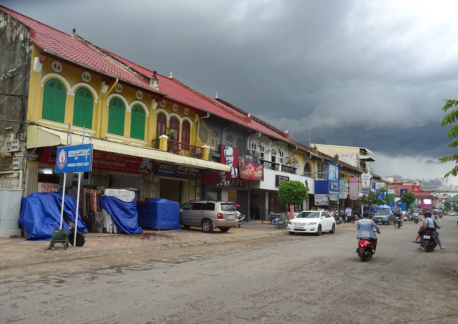 A terrace of yellow colonial shophouses in Battambang with green shuttered arched windows under a red tiled roof, storm cloud behind