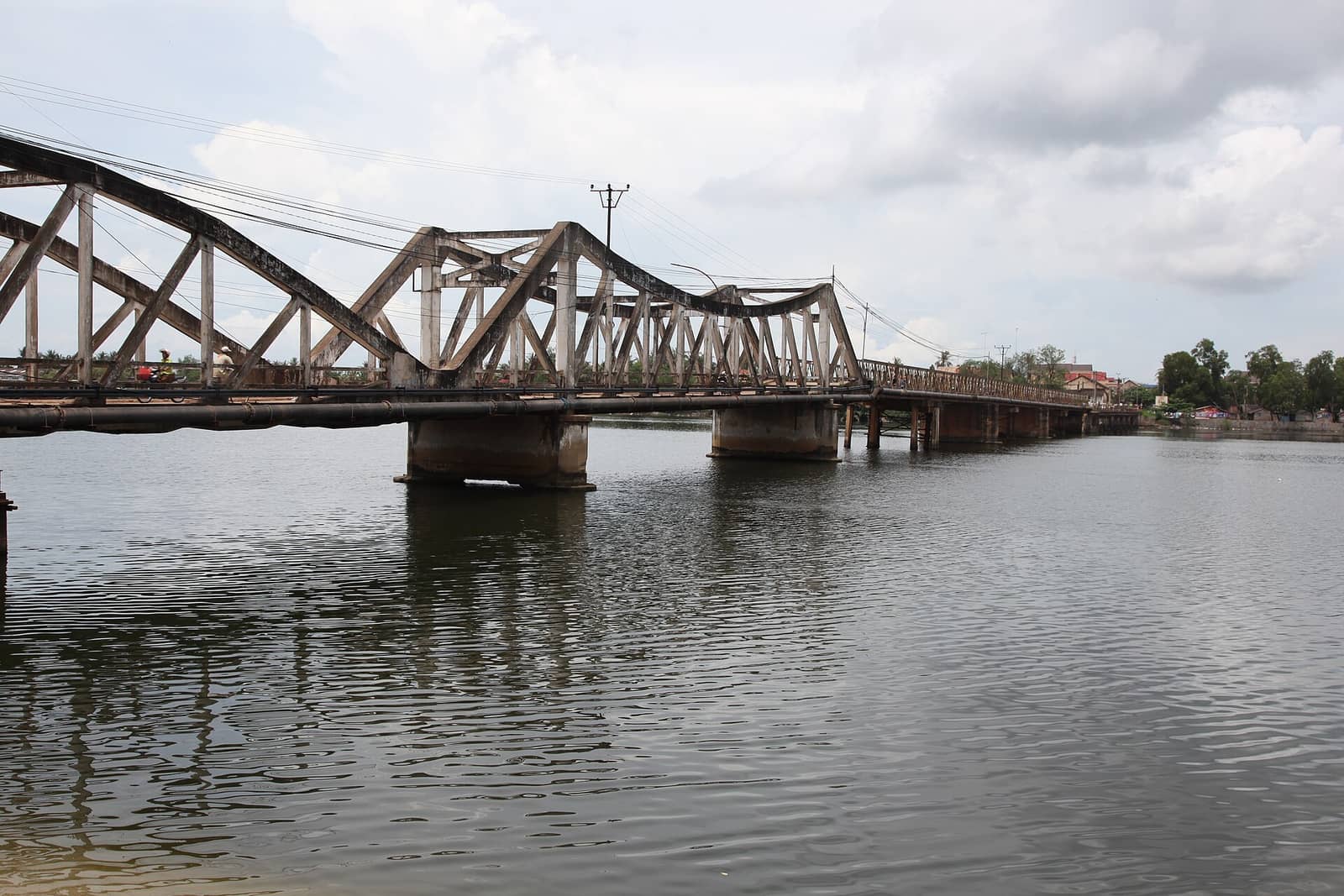 The old steel truss bridge across the Kampot River, its spans reflected in flat water with the town along the far bank