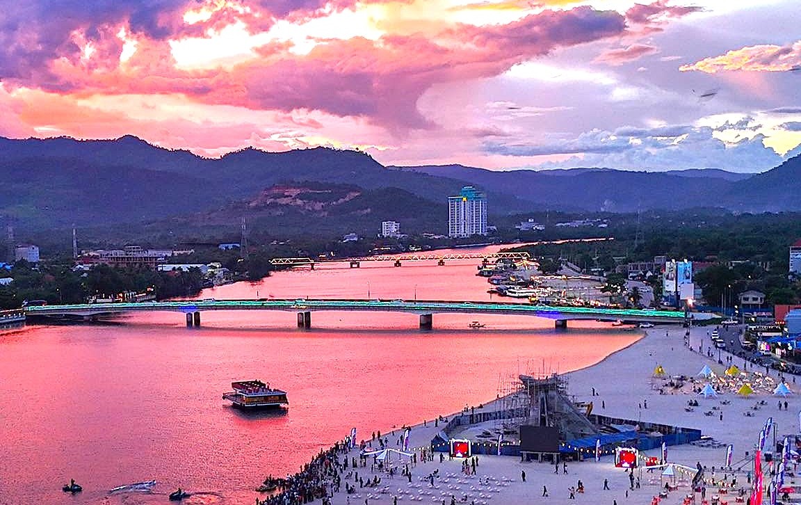 Sunset over the Kampot riverfront — the town beach busy with people, the lit road bridge and the older truss bridge beyond it, and the Bokor range on the skyline