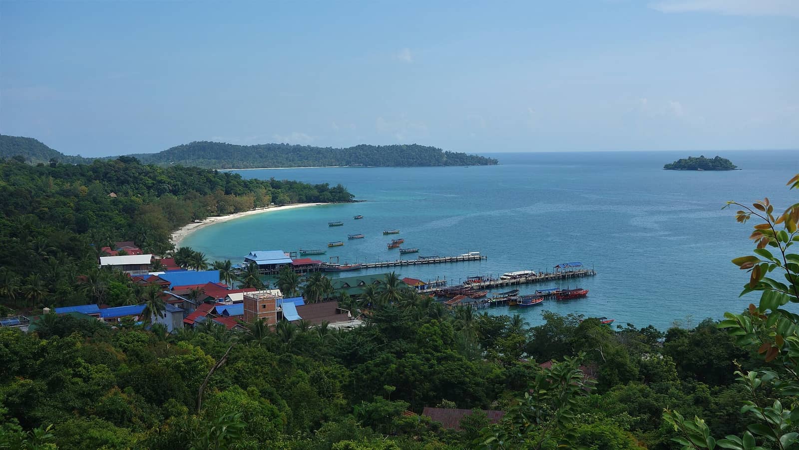 Koh Touch village on Koh Rong seen from the hillside above — red and blue roofs in the trees, a long pier lined with wooden boats, and a pale turquoise bay running out to a small island