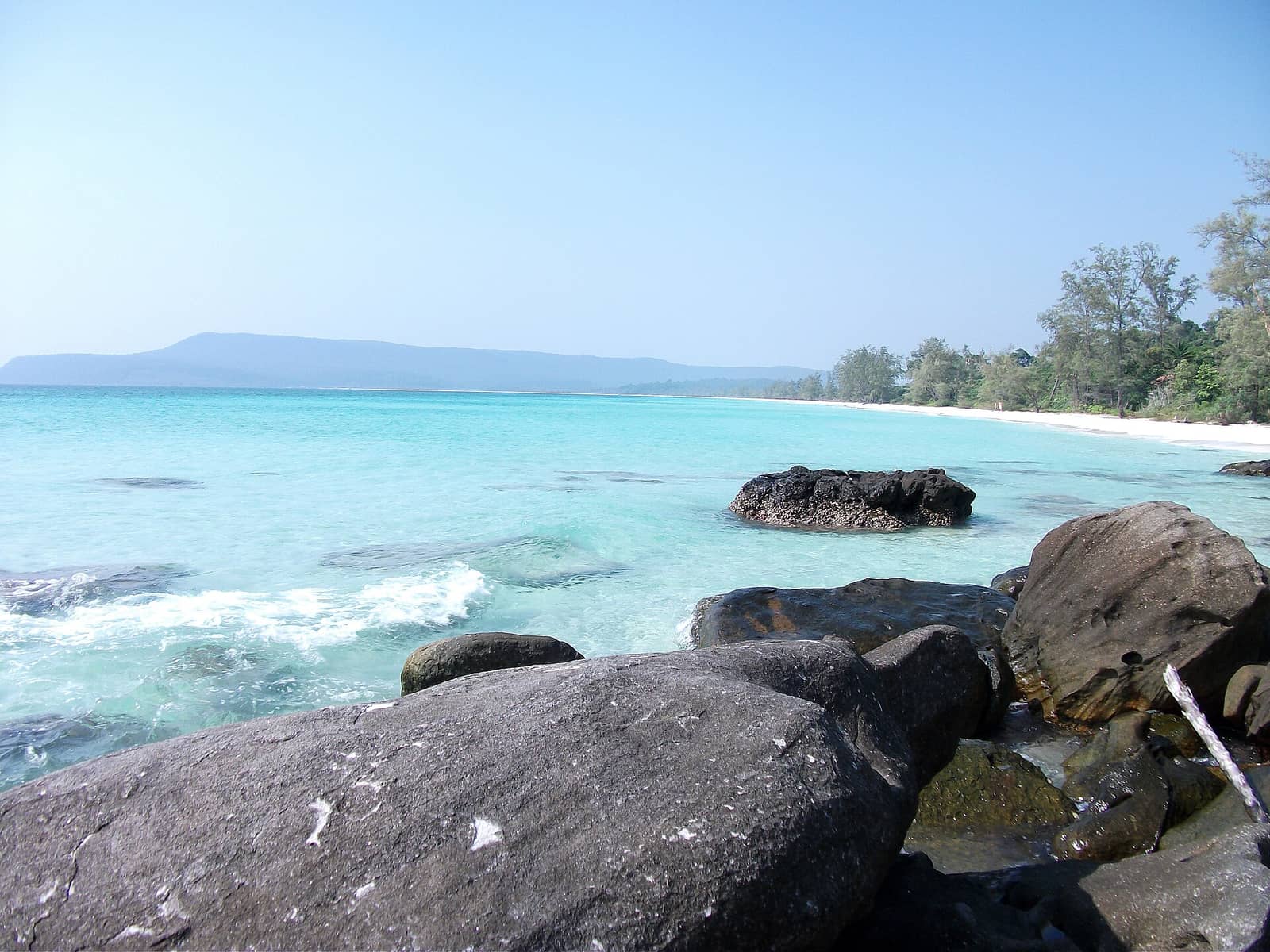 A wide curve of white sand and pale turquoise shallows on Koh Rong, dark boulders in the foreground and forested hills running away along the shore
