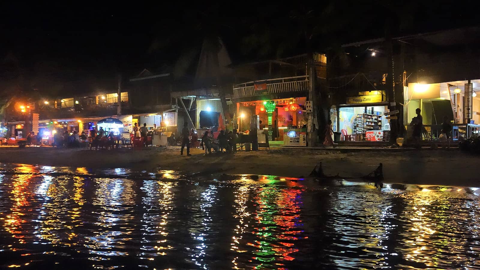 The beachfront strip at Koh Touch after dark, bar and restaurant lights along the sand thrown in coloured streaks across the shallows