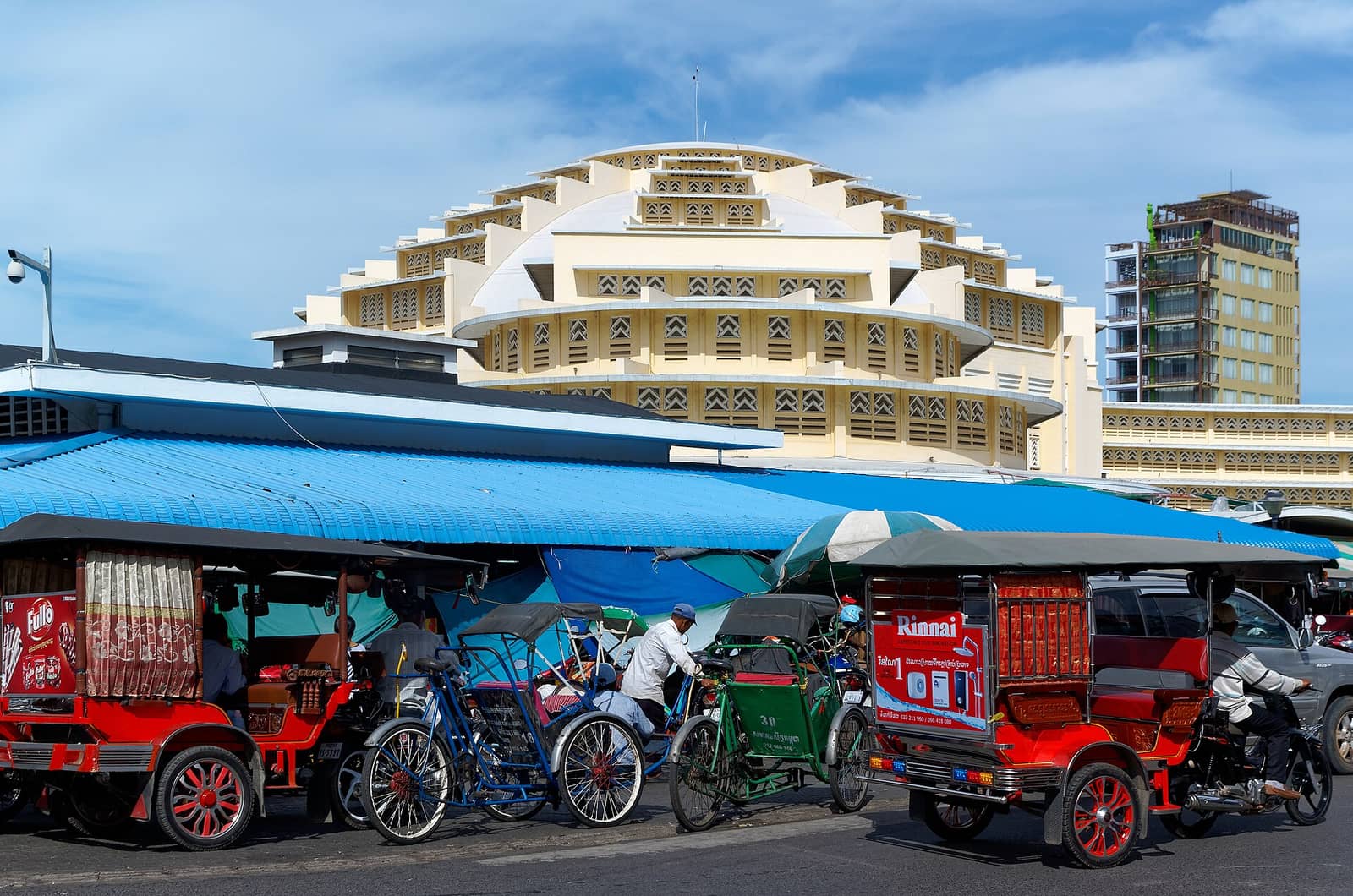 The pale yellow art deco dome of Phnom Penh’s Central Market behind a rank of cyclos and tuk-tuks waiting under blue awnings