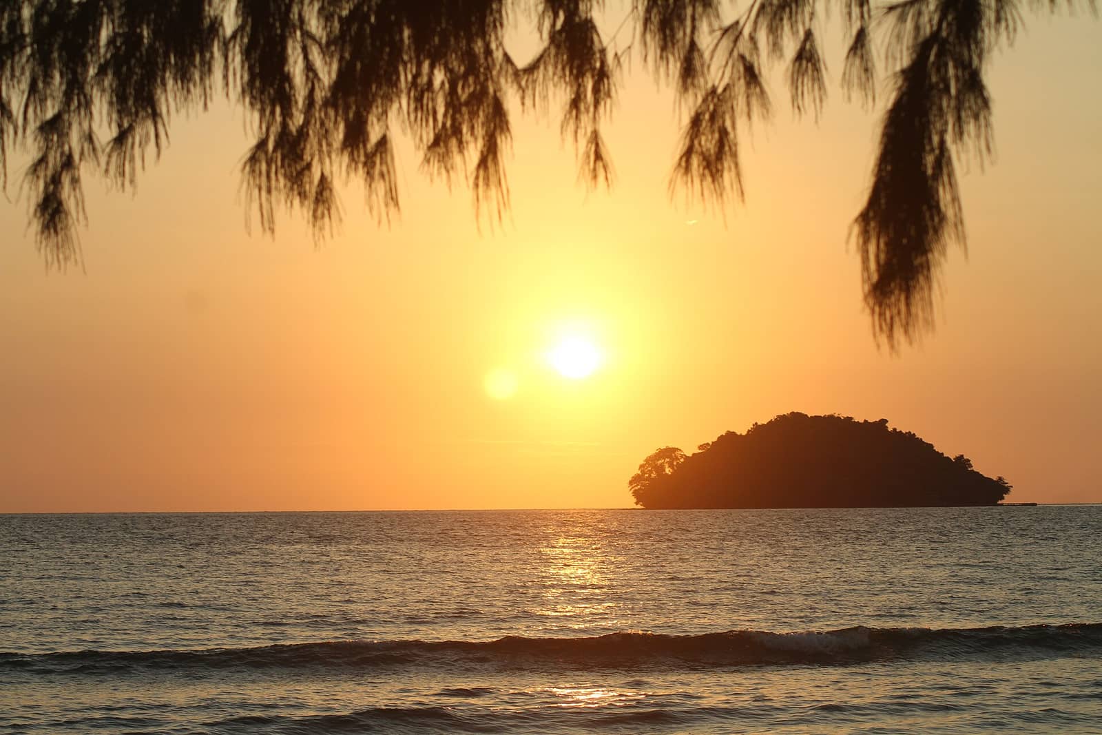 Sunset over a small forested island off Otres Beach, seen under the branches of a pine on the shore