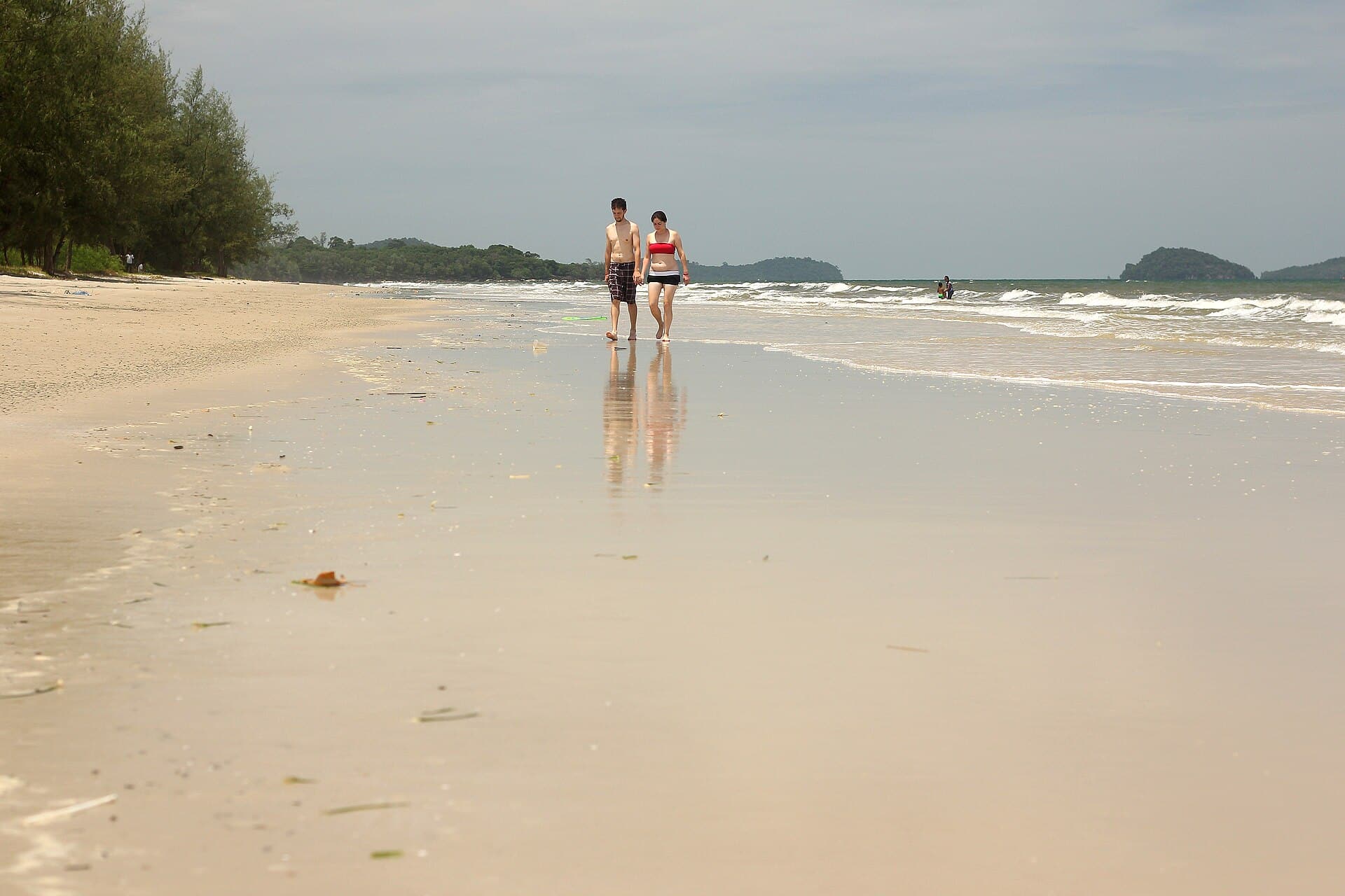 Two people walking the wet sand at the water’s edge on Otres Beach, Sihanoukville, their reflections under them and a forested headland out at the point